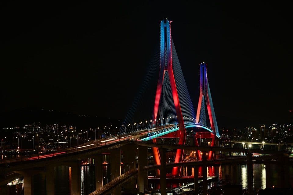 Un grande ponte strallato illuminato da luci rosse e blu attraversa l'acqua di notte, con uno skyline cittadino sullo sfondo.