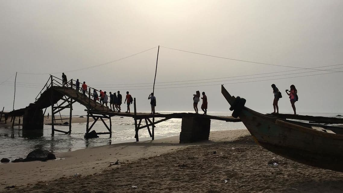 Un gruppo WeRoad in silhouette attraversa un ponte di legno rustico sull'acqua, partendo da una spiaggia sabbiosa.