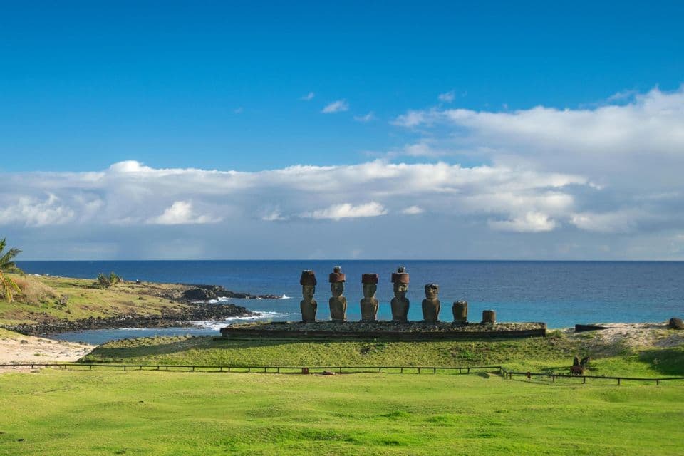 Una fila di grandi statue di pietra con chignon si erge su una piattaforma lungo una costa erbosa, affacciata sull'oceano sotto un cielo blu con nuvole bianche.