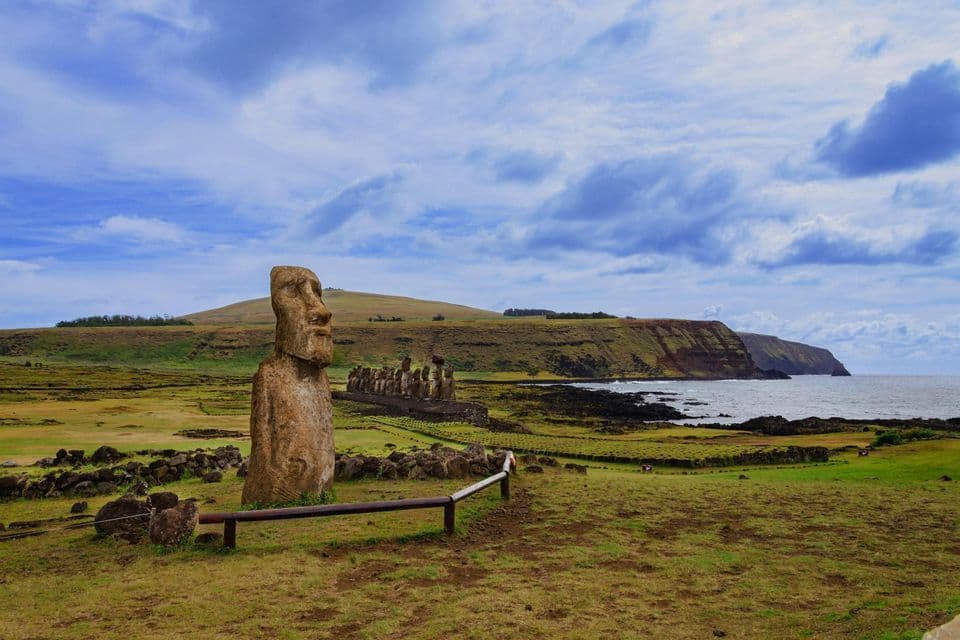 In primo piano, una statua Moai si erge in un campo erboso, con altre statue su una piattaforma in riva al mare sotto un cielo nuvoloso.