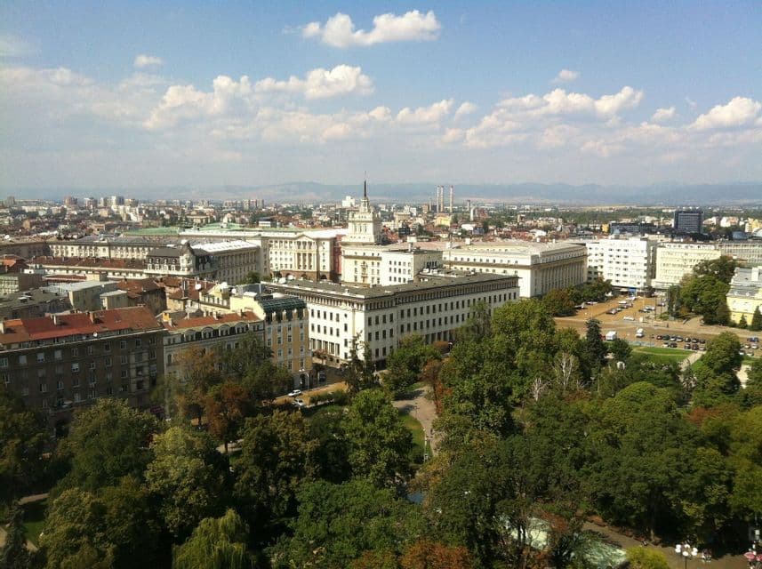 Una vista aerea di una città europea caratterizzata da grandi edifici storici bianchi, un lussureggiante parco verde e montagne lontane sotto un cielo blu.