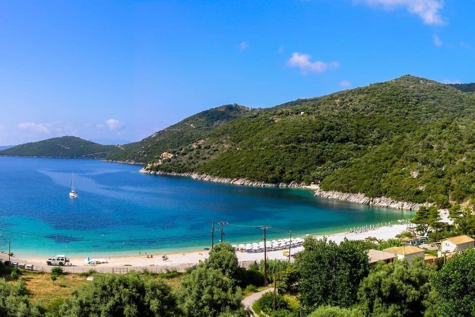 Ein Panoramablick auf einen Sandstrand in einer türkisfarbenen Bucht, umgeben von grünen Hügeln. Ein einzelnes Segelboot schwimmt auf dem ruhigen Wasser.