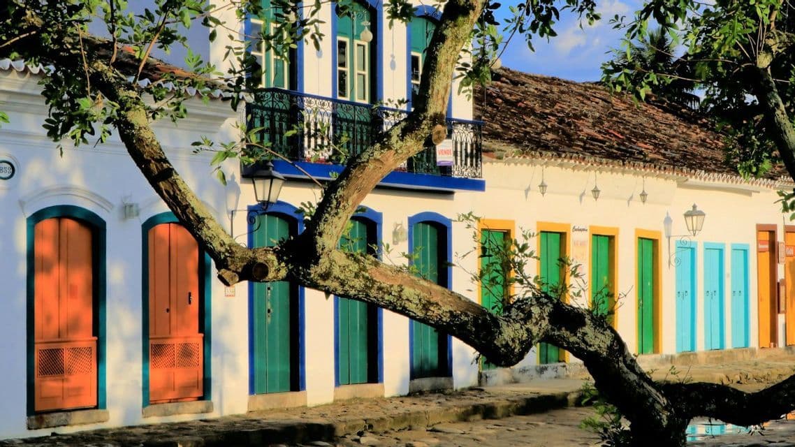 Ein moosbewachsener Ast im Vordergrund rahmt eine Reihe weißer Kolonialgebäude mit bunten Türen an einer Kopfsteinpflasterstraße ein.
