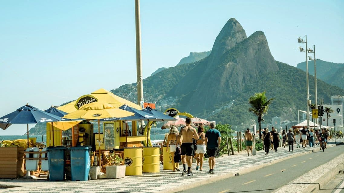 Menschen spazieren auf einer sonnigen, gefliesten Promenade, neben einem gelben Strandkiosk, mit einem großen, grünen Berg im Hintergrund.