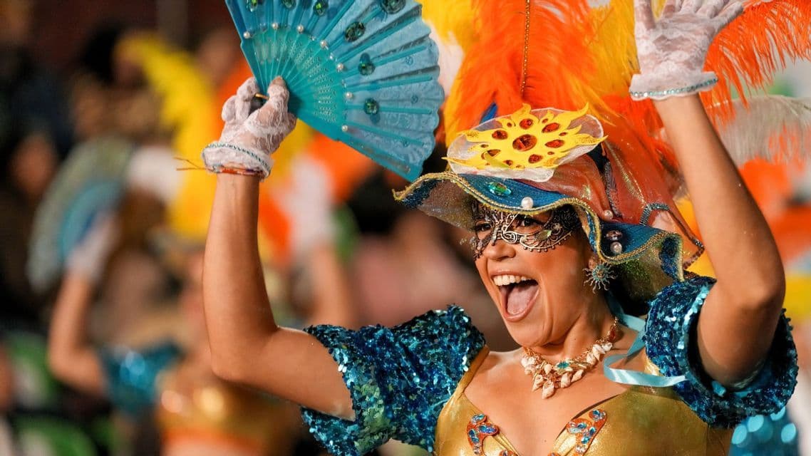 Eine lächelnde Frau tanzt in einem farbenfrohen Karnevalskostüm mit Maske, Federkopfschmuck und blauem Fächer.