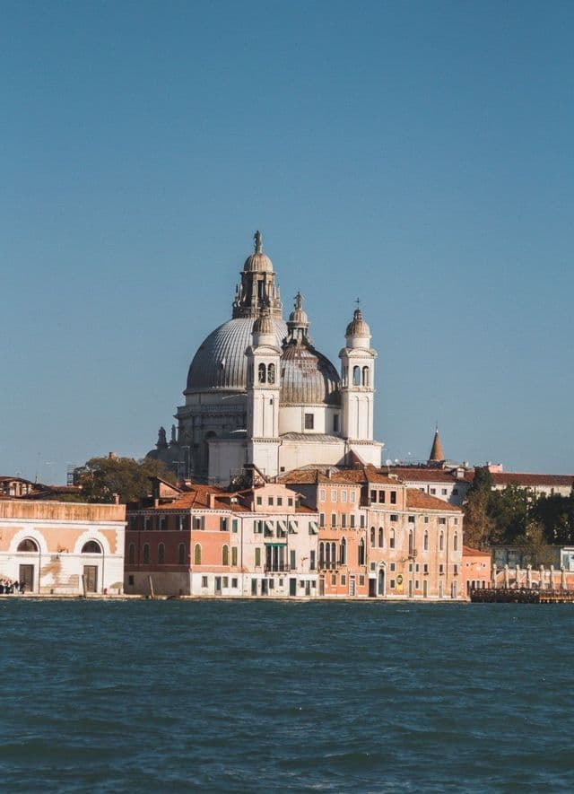 Una grande basilica a cupola bianca si erge sopra edifici colorati sul lungomare sotto un cielo azzurro e limpido.