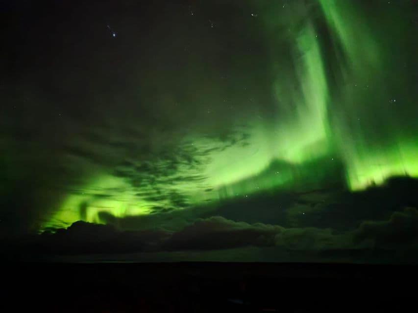 L'aurora boreale verde danza nel cielo notturno, sopra uno strato di nuvole e un orizzonte scuro, con stelle visibili sullo sfondo.