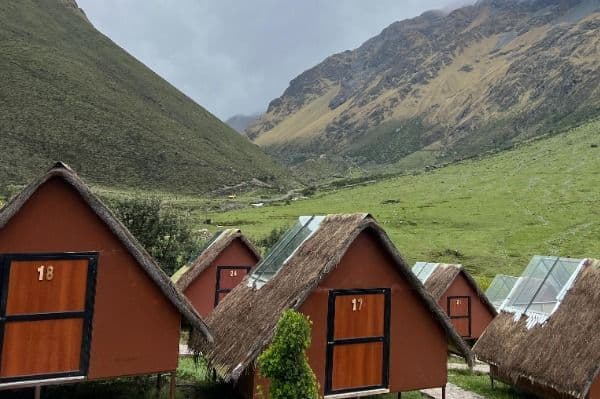Cabañas rústicas en forma de A, de color marrón rojizo, con techos de paja y claraboyas, situadas en un exuberante valle de montaña verde bajo un cielo nublado.