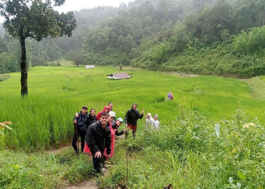 Un viaje en grupo de WeRoad con equipo de lluvia posa en un sendero junto a exuberantes arrozales verdes con una colina boscosa al fondo.