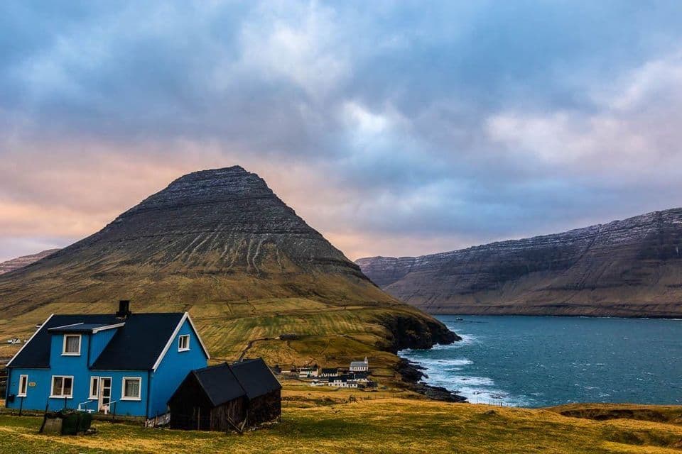 Una casa azzurra brillante si trova su una collina erbosa che si affaccia su un piccolo villaggio costiero ai piedi di una grande montagna sotto un cielo nuvoloso.