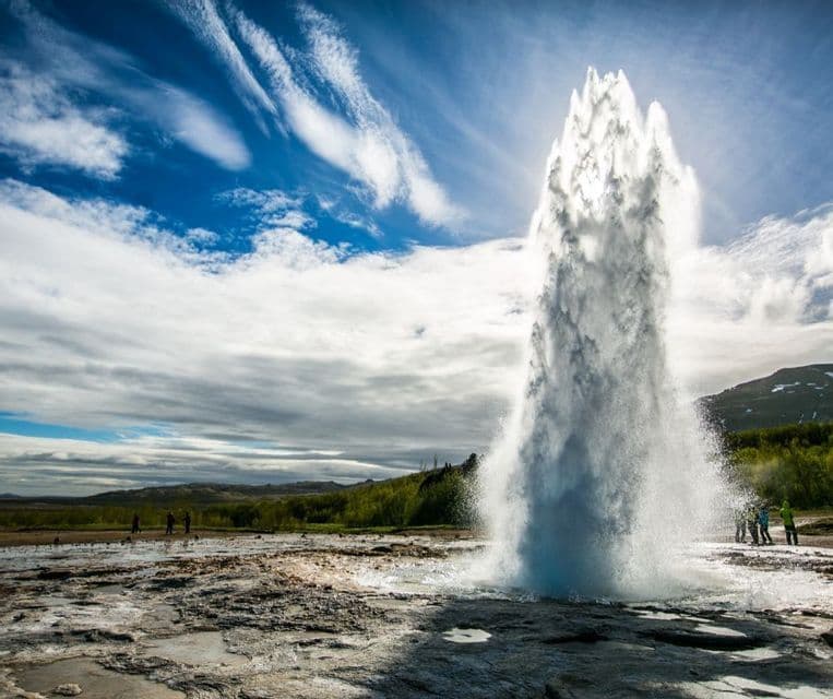 Un alto geyser erutta dal terreno roccioso, spruzzando acqua in aria sotto un cielo drammatico blu e bianco, mentre le persone lo osservano.