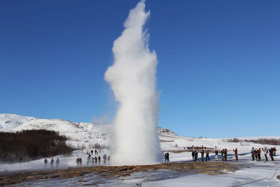 Durante un viaggio di gruppo WeRoad, si osserva un grande geyser eruttare, proiettando un'alta colonna di vapore nel cielo azzurro sopra un paesaggio innevato.