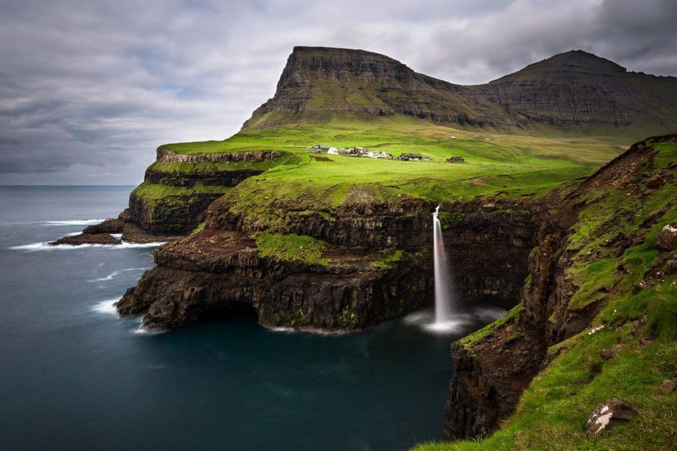 Una cascata si getta da una verde scogliera erbosa direttamente nell'oceano, con un piccolo villaggio e montagne sullo sfondo sotto un cielo coperto.