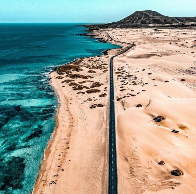 Veduta aerea di una lunga strada che si snoda tra un deserto di sabbia con dune e una costa oceanica turchese, con una montagna sullo sfondo.