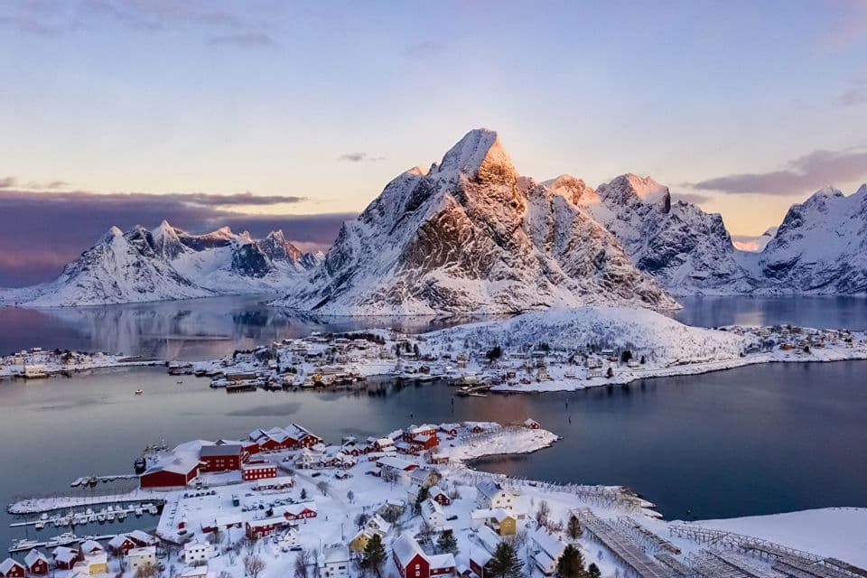 Eine Luftaufnahme eines verschneiten Küstendorfes mit roten Häusern, gelegen am Fuße großer, schneebedeckter Berge bei Sonnenaufgang.