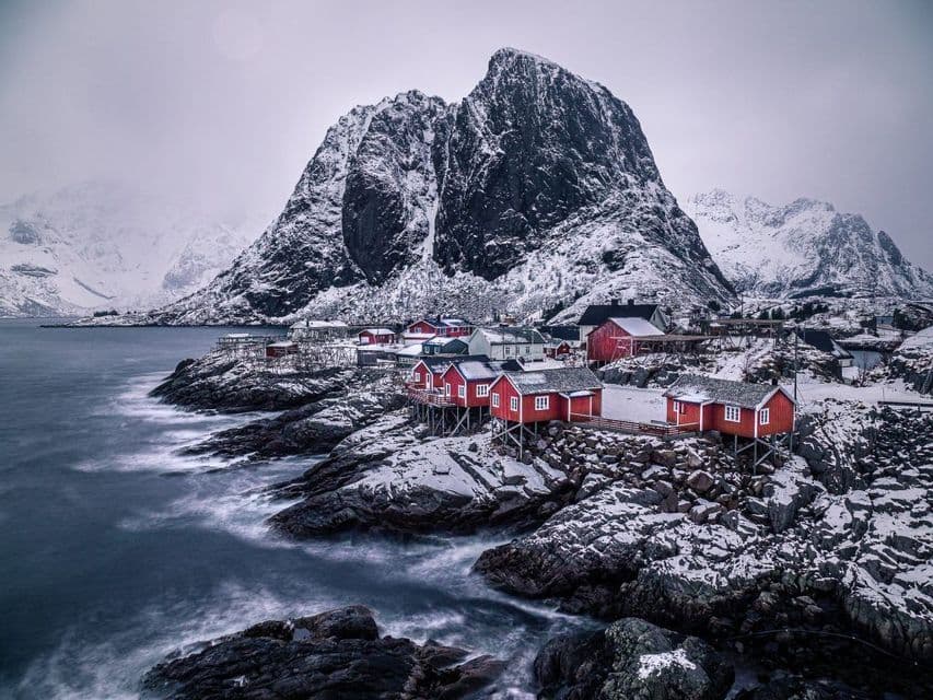 Des maisons en bois rouges sur pilotis bordent une côte enneigée et rocheuse au pied d'une grande montagne, avec les vagues qui s'écrasent sur le rivage.