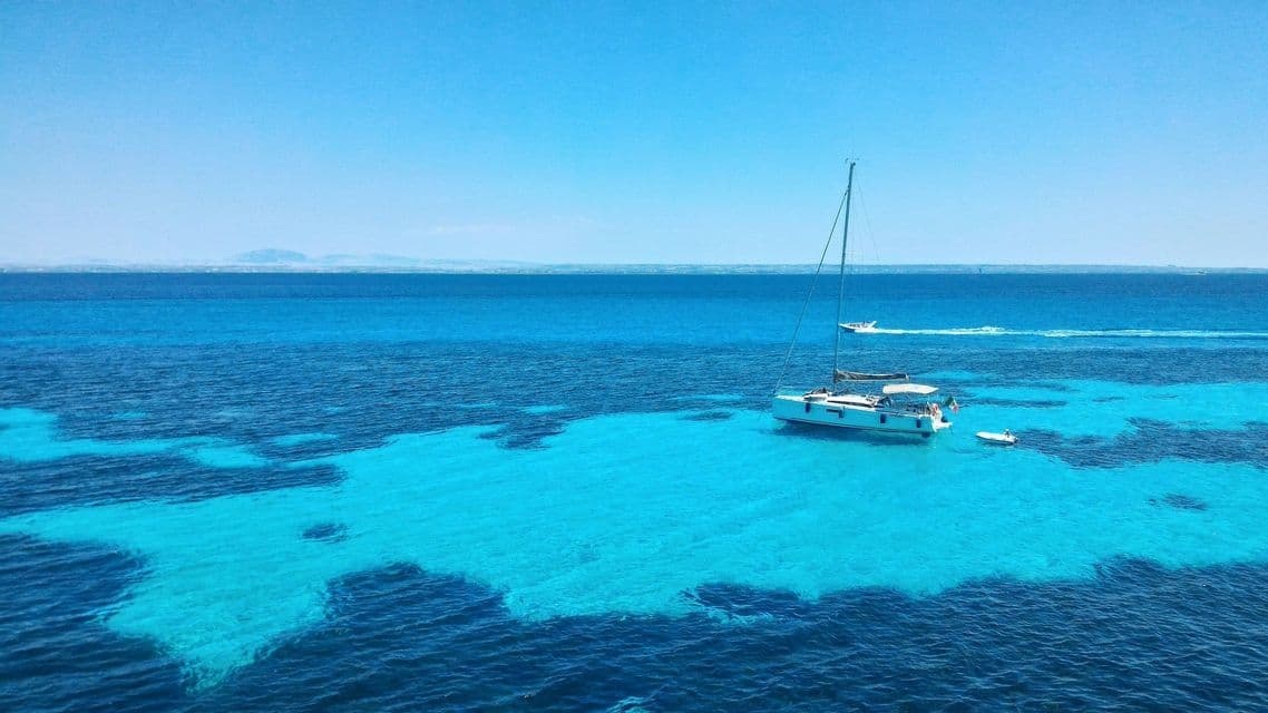 Una barca a vela bianca ancorata in acque turchesi vibranti, in contrasto con il mare blu profondo sotto un cielo sereno.