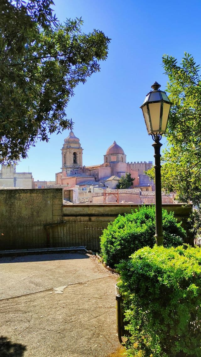 Una vista di una città storica con una cattedrale imponente, incorniciata da alberi verdi e un lampione classico sotto un cielo azzurro.