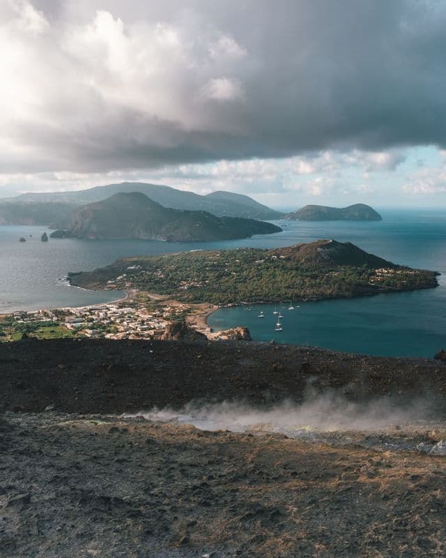 Una vista da un pendio vulcanico fumante che domina una città costiera su un'isola lussureggiante, circondata dal mare e da altre isole.