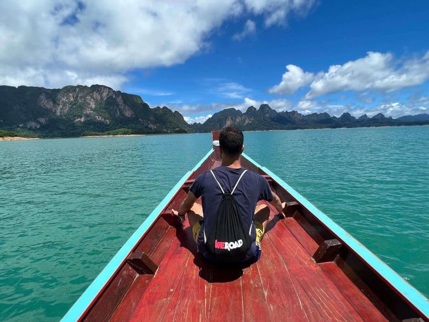 Une personne avec un sac à dos WeRoad est assise à la proue d'un bateau en bois, regardant un lac turquoise vers des montagnes lointaines.
