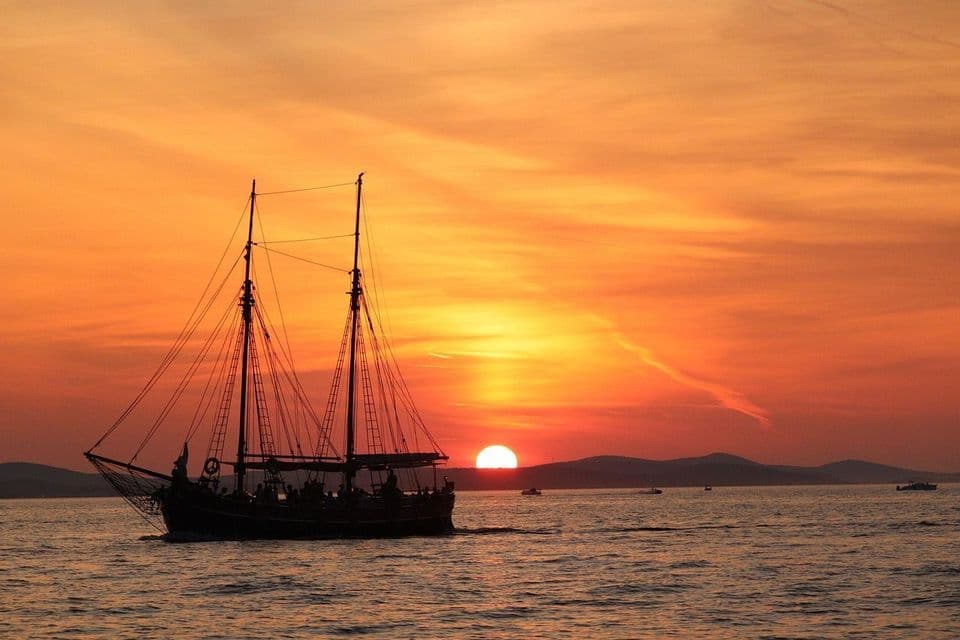 A silhouetted two-masted sailing ship carrying a WeRoad group trip sails on the ocean during a vibrant orange sunset.