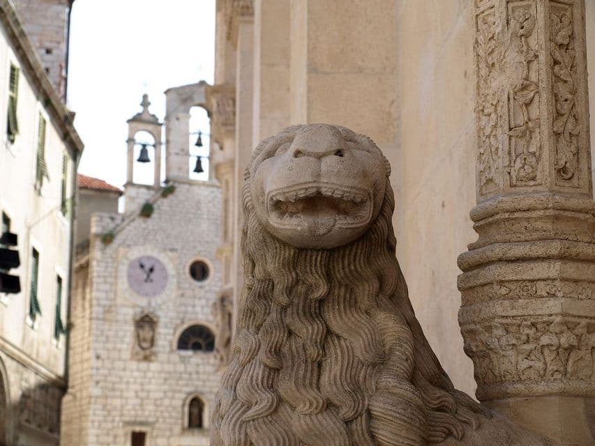 A carved stone lion statue with an open mouth stands guard next to a pillar, with a historic stone clock tower in the background.