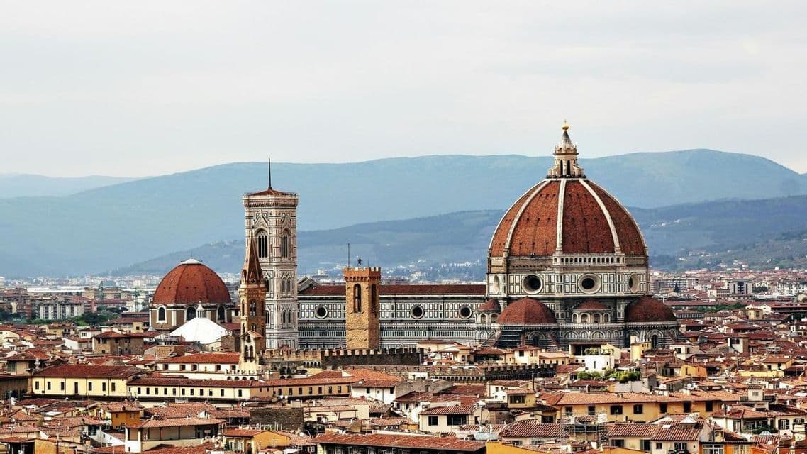 La gran cúpula de tejas rojas y el campanario de una catedral se alzan sobre los tejados de terracota de una ciudad, con montañas visibles al fondo.