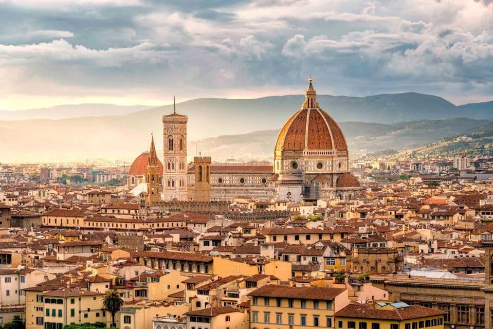 Vista panorámica de una ciudad mostrando una catedral histórica con una gran cúpula de terracota y campanario, con montañas distantes bajo un cielo nublado.