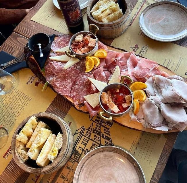 Un plato de madera con embutidos, queso, rodajas de naranja y pan servido con una botella de vino en la mesa de un restaurante.
