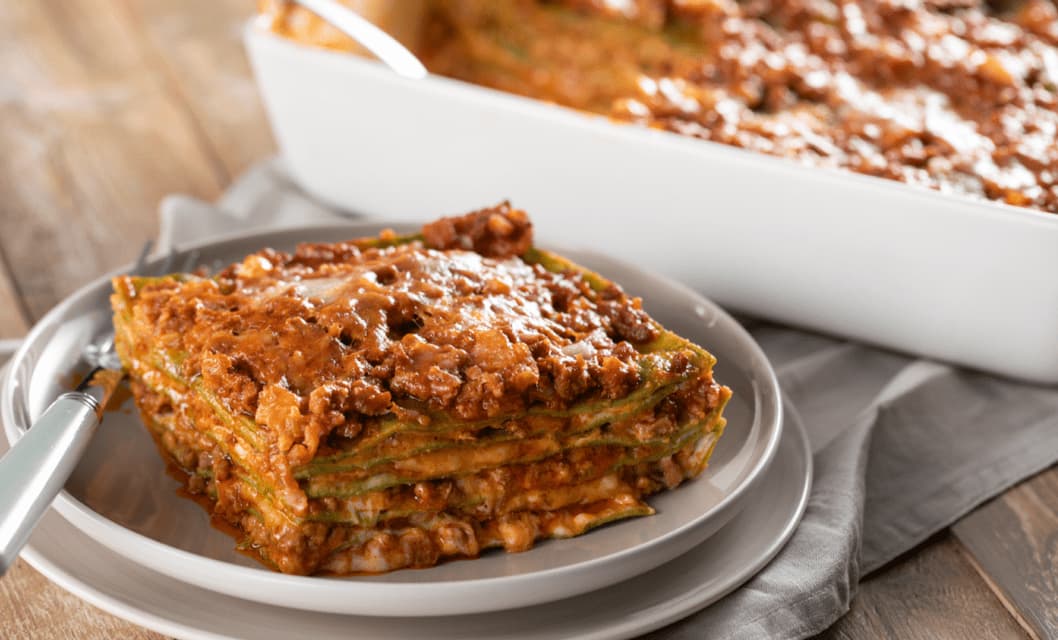 Una porción cuadrada de lasaña verde con salsa de carne en un plato gris, con una fuente para hornear blanca de fondo sobre una mesa de madera.