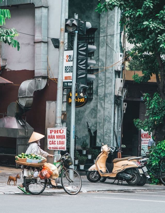 Eine Person mit einem Kegelhut schiebt ein mit einem Korb grüner Früchte beladenes Fahrrad entlang eines städtischen Gehwegs neben einem geparkten Motorroller.