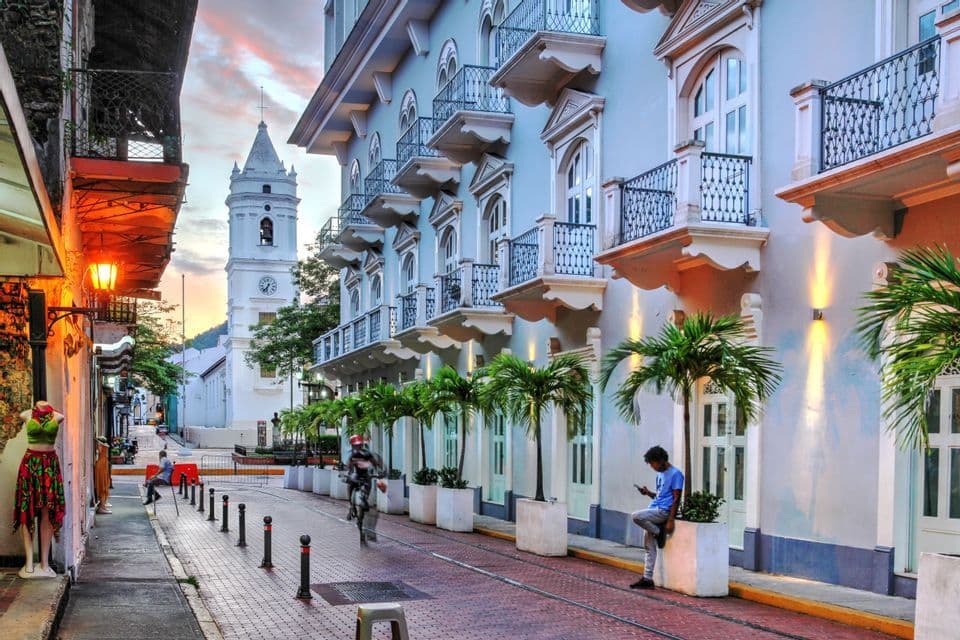 Una stretta strada di mattoni fiancheggiata da edifici coloniali e palme al tramonto, con una torre dell'orologio bianca in lontananza.