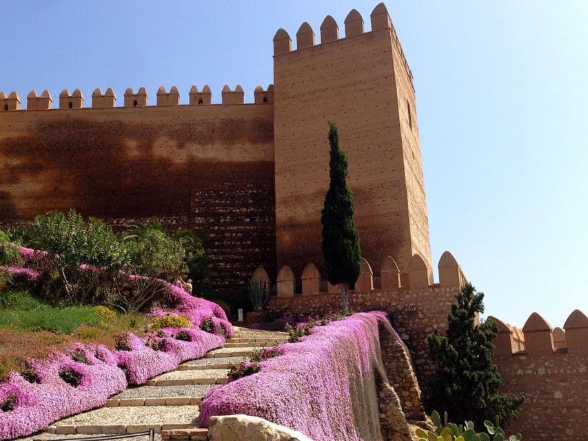 Escalones de piedra ascienden una colina tapizada de flores rosadas en cascada, llevando a las murallas almenadas de una antigua fortaleza bajo un cielo despejado.