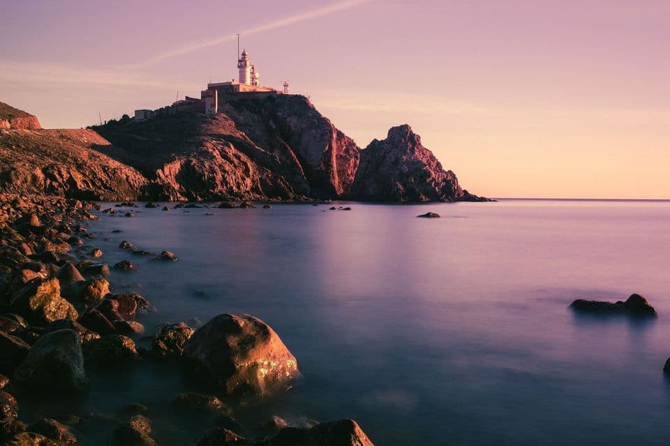 Un faro blanco se asienta sobre un acantilado rocoso con vistas a un mar en calma y una costa pedregosa al atardecer.