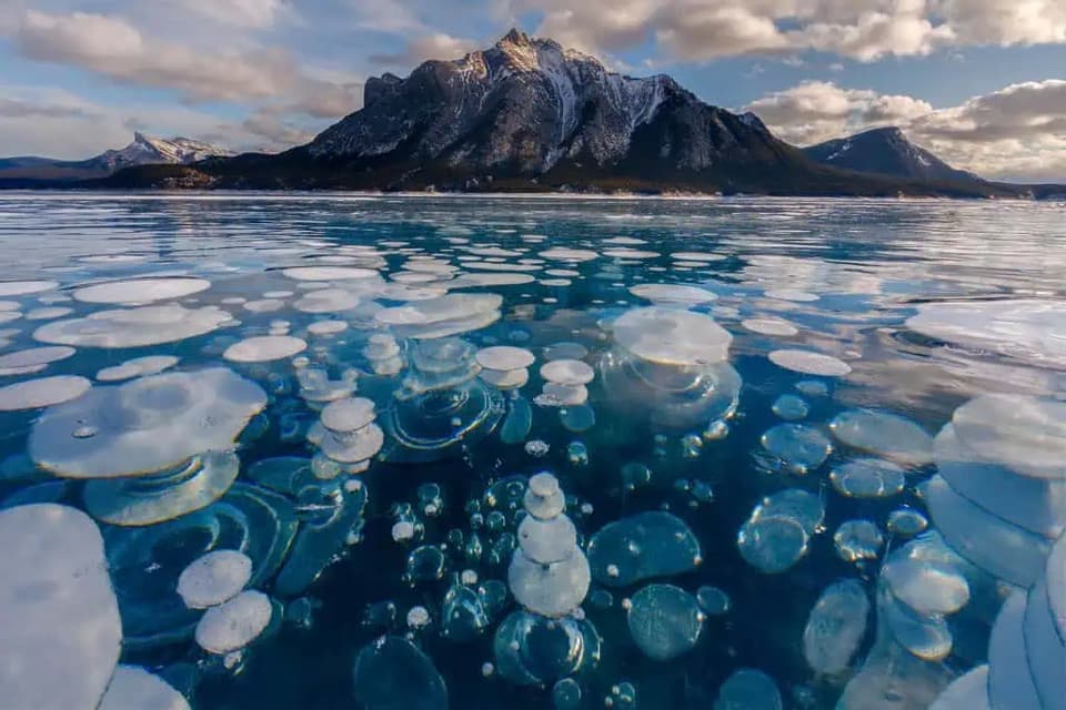 Bolle bianche ghiacciate sono intrappolate sotto il ghiaccio trasparente di un lago, con un'imponente montagna innevata sullo sfondo.