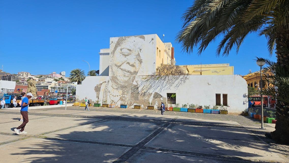 Ein großes Wandgemälde eines lächelnden Frauengesichts ziert ein Gebäude an einem sonnigen Stadtplatz, mit vorbeigehenden Passanten und einer Palme im Vordergrund.