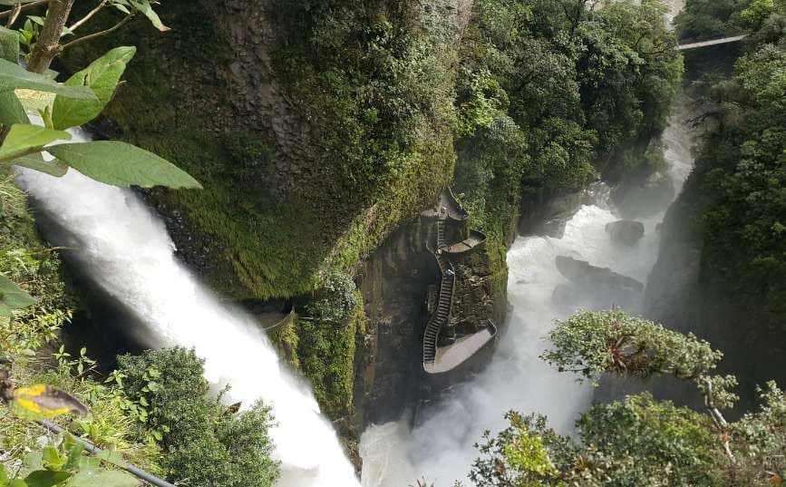 A view from above of a large waterfall cascading into a lush green gorge, with a winding stone staircase on the cliffside.