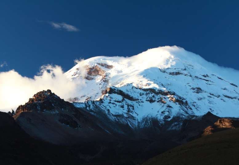 Sunlight illuminates a snow-covered mountain peak, with its base in shadow against a deep blue sky.