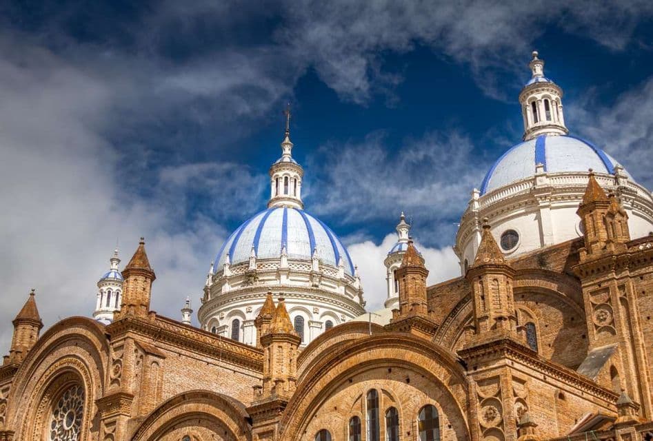 A low-angle view of an ornate brick cathedral with large blue and white striped domes against a cloudy sky.
