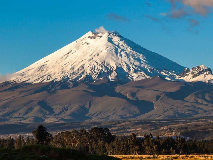 Un gran volcán cubierto de nieve emite una columna de humo desde su cima, dominando un paisaje de colinas y árboles bajo un cielo azul claro.