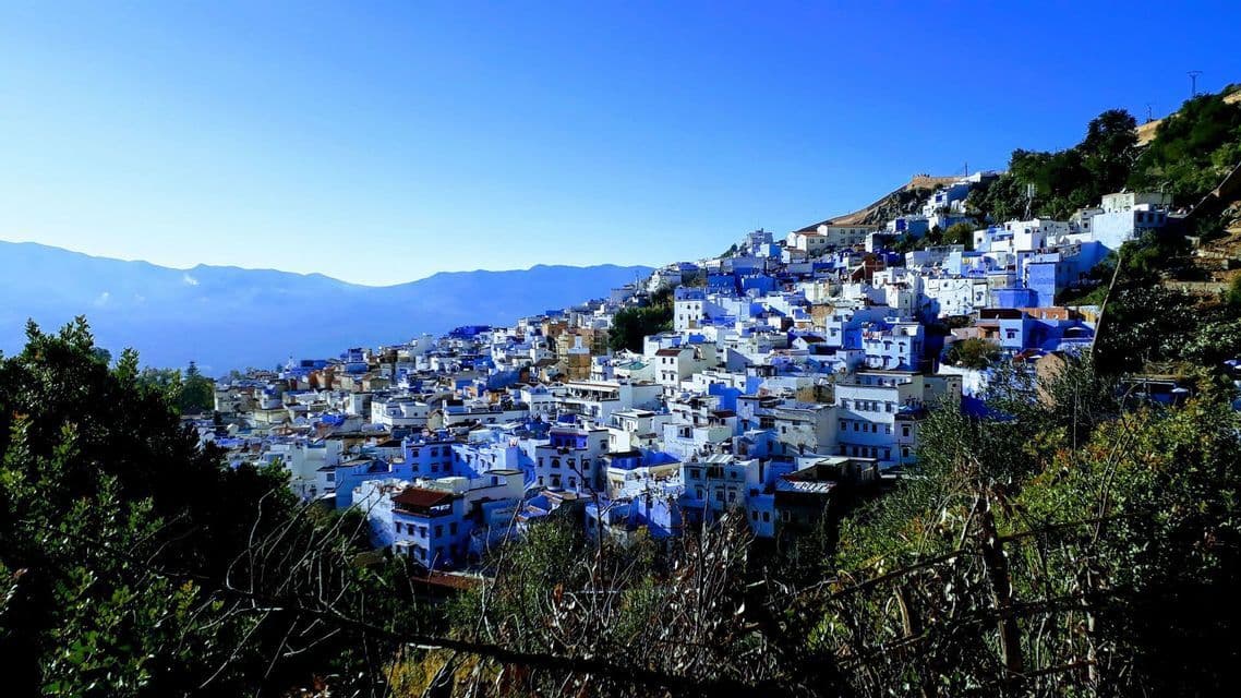 Una ciudad de edificios azules y blancos cubre una ladera verde, con montañas distantes bajo un cielo despejado.