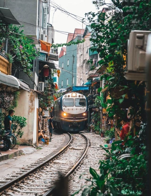 Un tren con el faro encendido viaja por las vías a través de una estrecha calle residencial bordeada de edificios y exuberante vegetación.