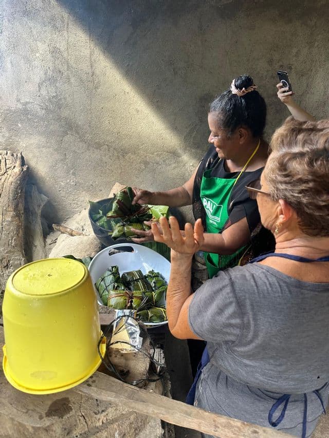 Une femme en tablier vert montre à une autre comment préparer des aliments enveloppés dans des feuilles lors d'un cours de cuisine de groupe WeRoad.