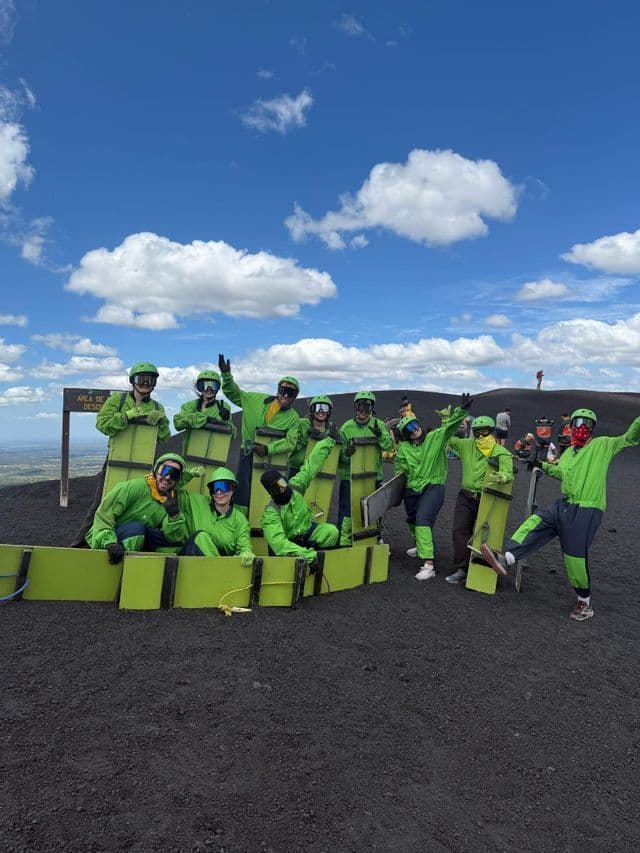 Un voyage de groupe WeRoad en combinaisons vert vif et lunettes posant avec des planches sur une sombre pente volcanique sous un ciel bleu.