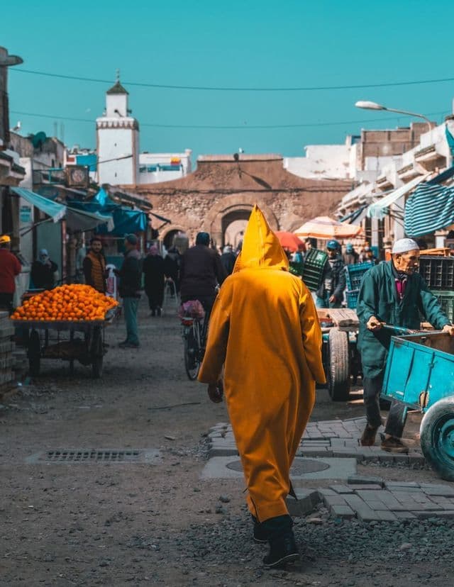Una persona con una túnica amarilla con capucha camina por una concurrida calle de mercado con un carro de naranjas y un minarete a lo lejos.