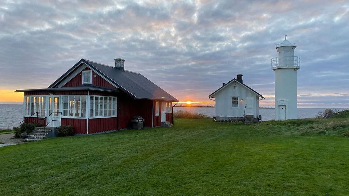 Ein rotes Haus und ein weißer Leuchtturm stehen auf einer grünen Wiese mit Meerblick, während die Sonne am Horizont untergeht.