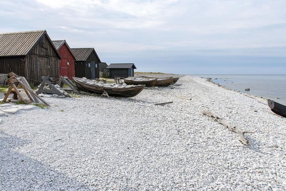 Alte Holzboote liegen an einem weißen Kieselstrand vor rustikalen hölzernen Fischerhütten am Meer.