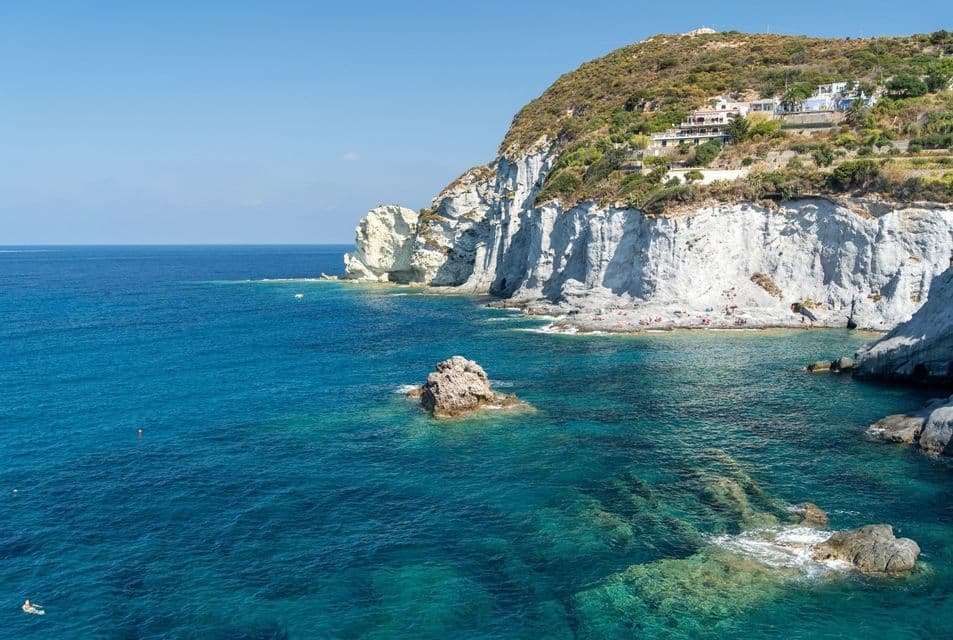 Vista dall'alto di un mare turchese contro scogliere bianche, con persone su una piccola spiaggia alla base e ville sulla verde collina sovrastante.