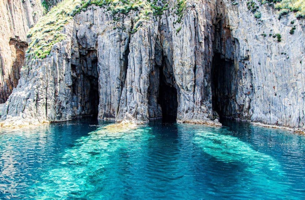 Le scure aperture di varie grotte marine sono scavate in una scogliera impervia, a picco su acque cristalline e turchesi, illuminate dal sole.