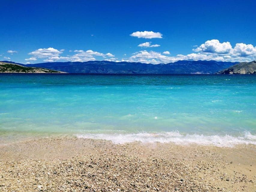 Una spiaggia di ciottoli bagnata da acque turchesi, con montagne in lontananza sotto un cielo azzurro intenso e nuvole bianche sparse.